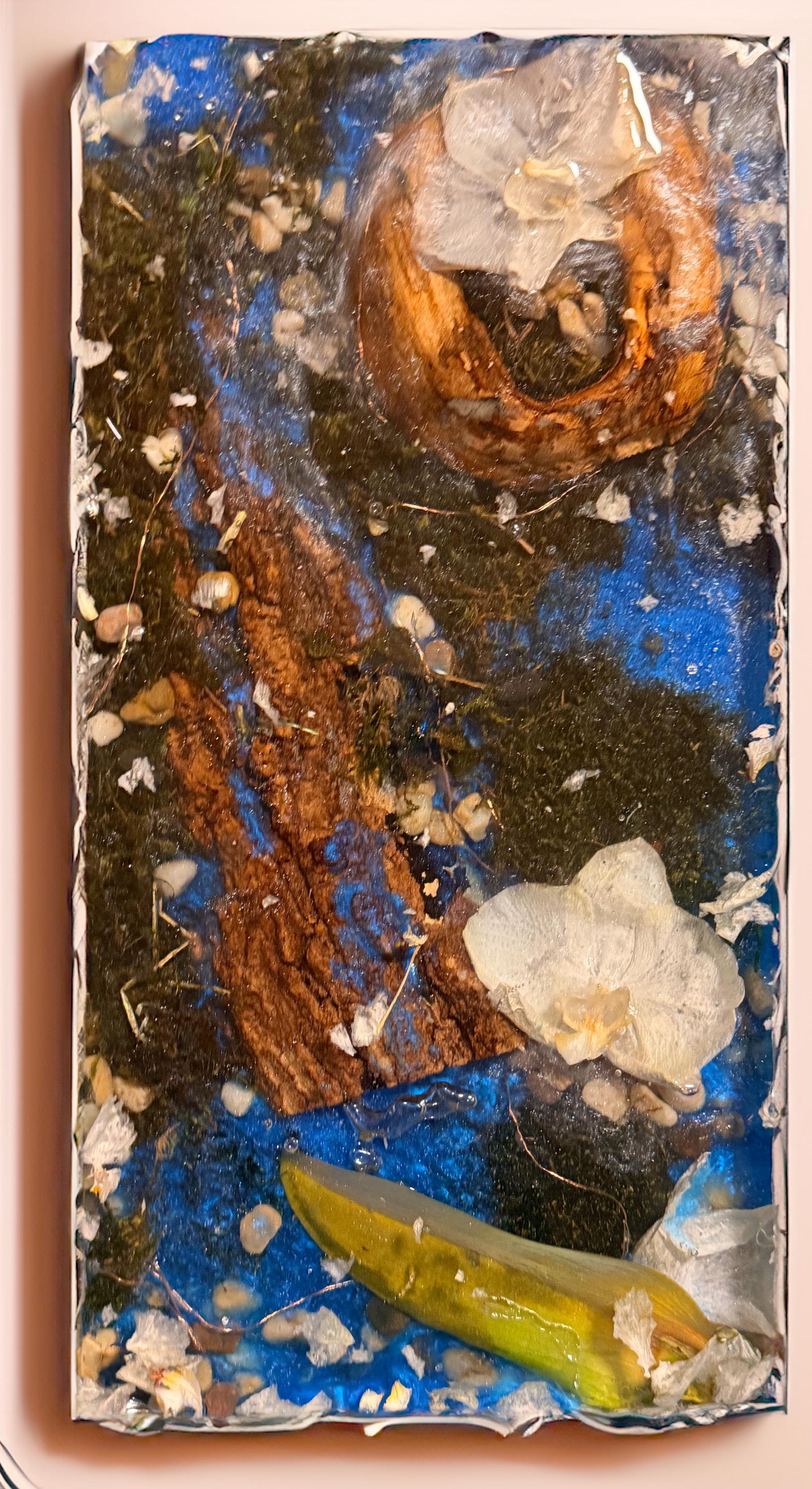 May include: Abstract art piece featuring a mix of natural elements encased in resin. The artwork includes a wooden ring, moss, small stones, and a yellow-green leaf against a blue background. The piece is framed in white.
